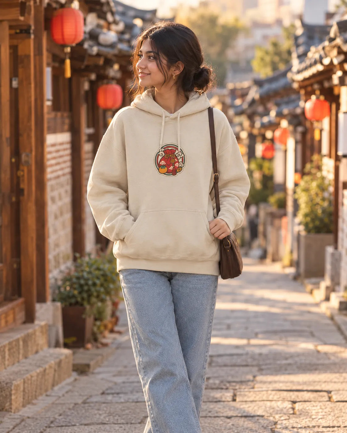 Woman walking down a lantern-lit Korean alley at golden hour, wearing a beige Luck Loading Korean hoodie with a small lucky pouch front chest print