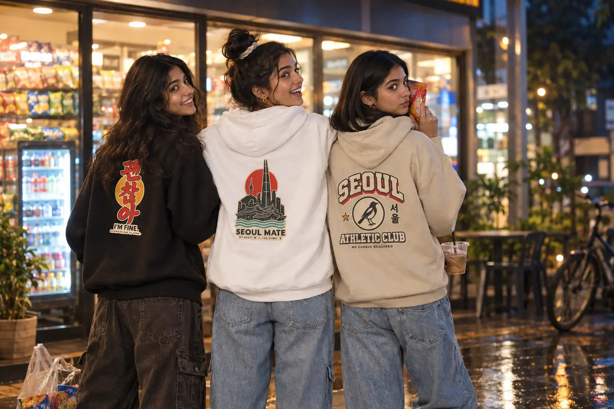 Three young women standing side-by-side in front of a brightly lit convenience store at night, showcasing matching hoodies with different graphic designs. The hoodie on the left is black with a red and yellow graphic, the center hoodie is white with a red and black graphic, and the right hoodie is beige with a red and black graphic. Three girls in Wear Pardesi Korean hoodies showing I'm Fine, Seoul Mate and Seoul Athletic Club back prints