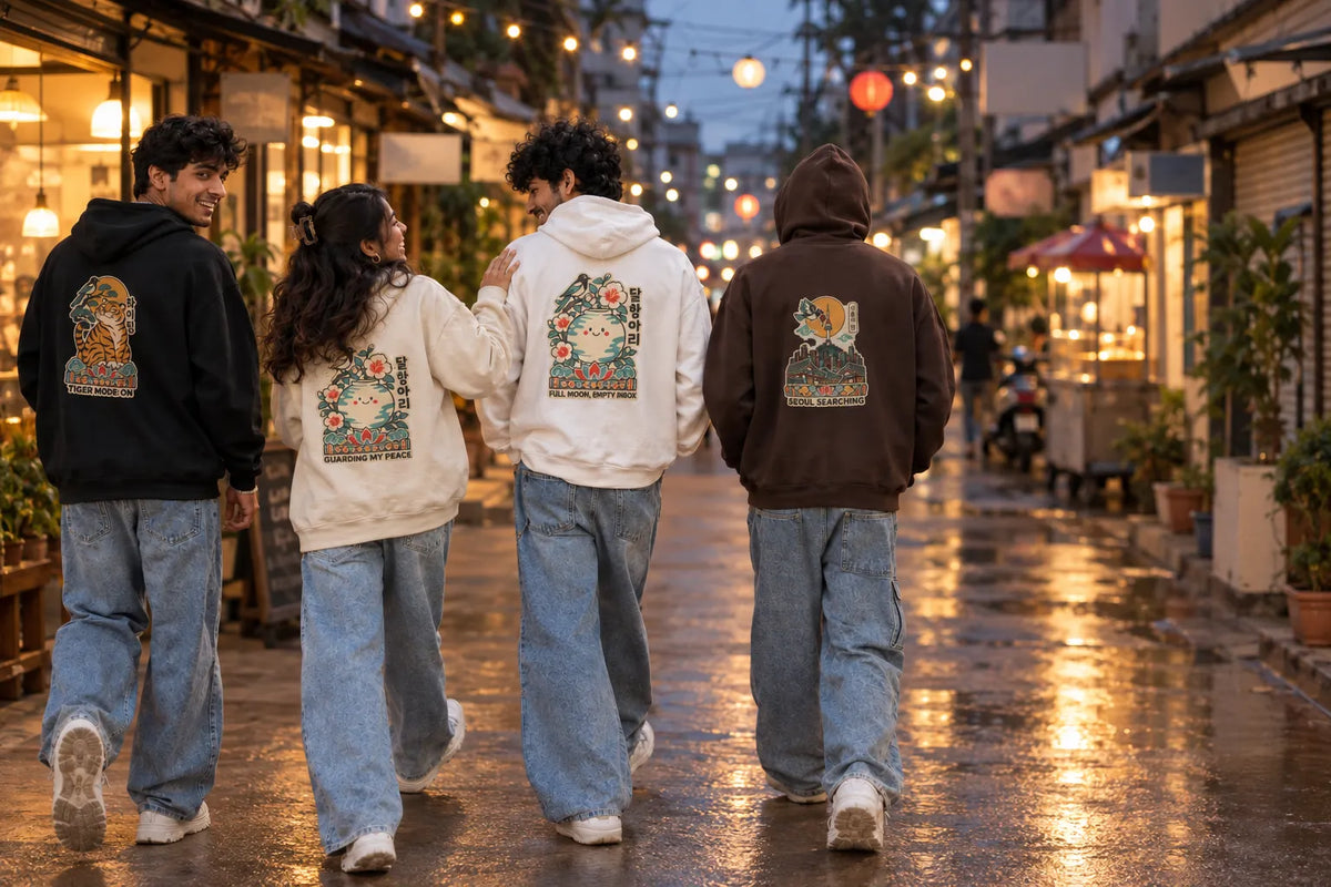 Four people walking away on a wet, warmly lit street, each wearing a different color hoodie with a large colorful graphic print on the back. The hoodies are black, cream, off-white, and dark brown, paired with wide-leg light blue jeans. Four friends walking down a lit-up street at night in Wear Pardesi Korean hoodies.