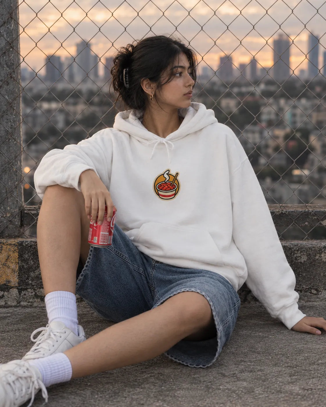 Woman sitting on a rooftop by a chain-link fence at sunset, wearing an off-white Eat First Think Later Korean hoodie with a small front chest print