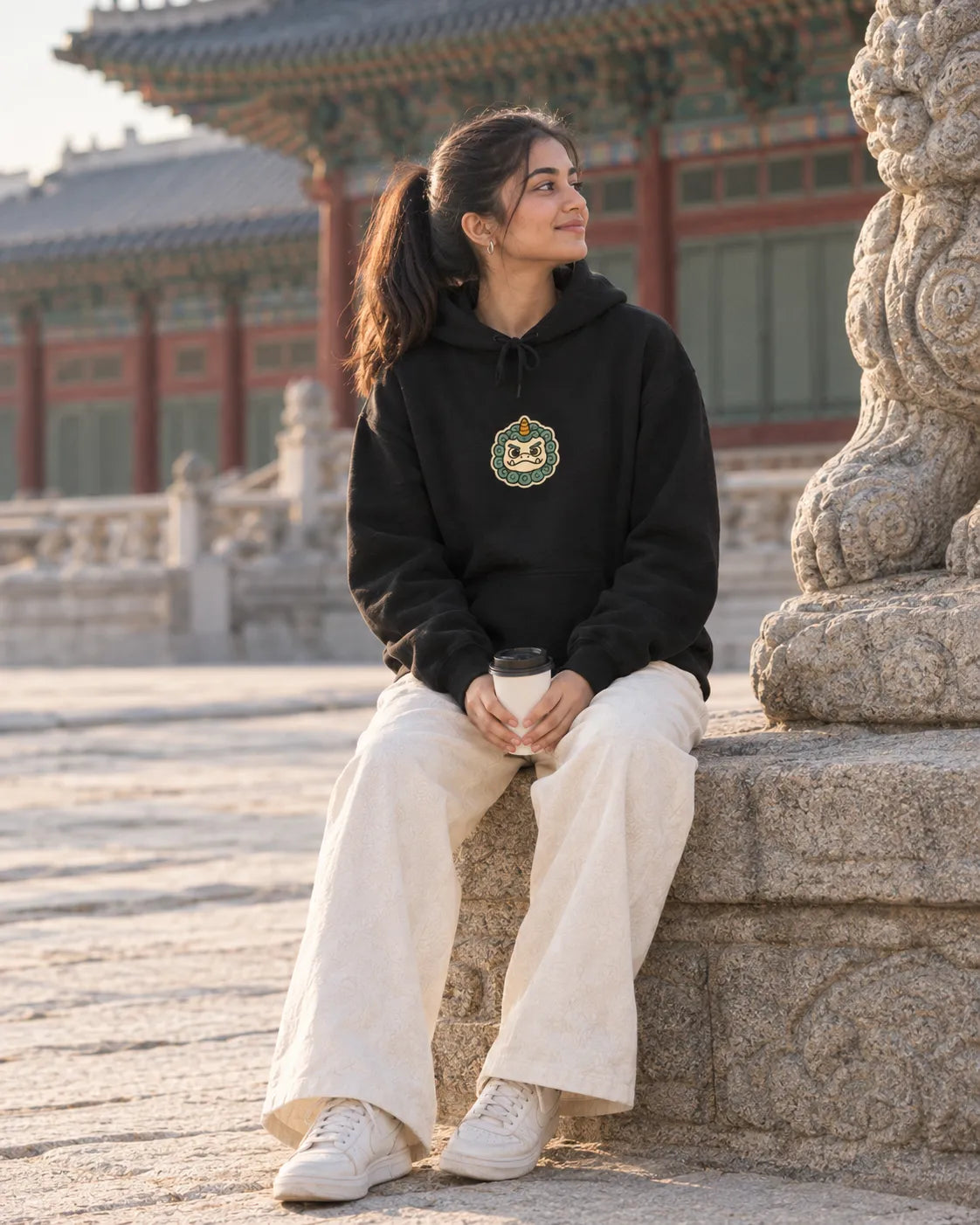 Woman sitting beside a stone guardian lion at a Korean palace, wearing a black Guarding My Peace Korean hoodie with a small front chest print