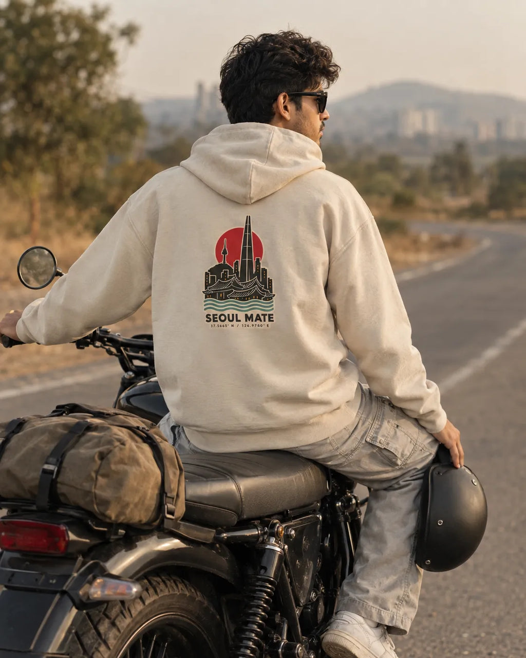 Man sitting on a black motorcycle on an open highway, wearing a beige Seoul Mate Korean hoodie with a Seoul skyline back print