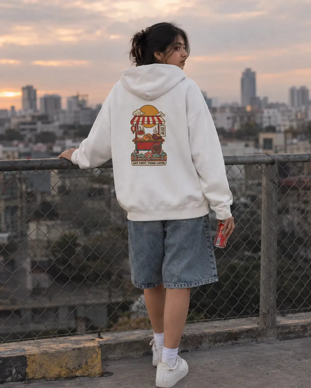 Woman on a city rooftop at sunset in an off-white Eat First Think Later Korean hoodie with denim shorts
