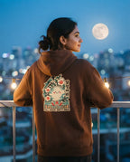 A woman stands on a balcony at dusk wearing a brown hoodie with a large colorful cat graphic and Korean text on the back, with a full moon and blurred city lights in the background.