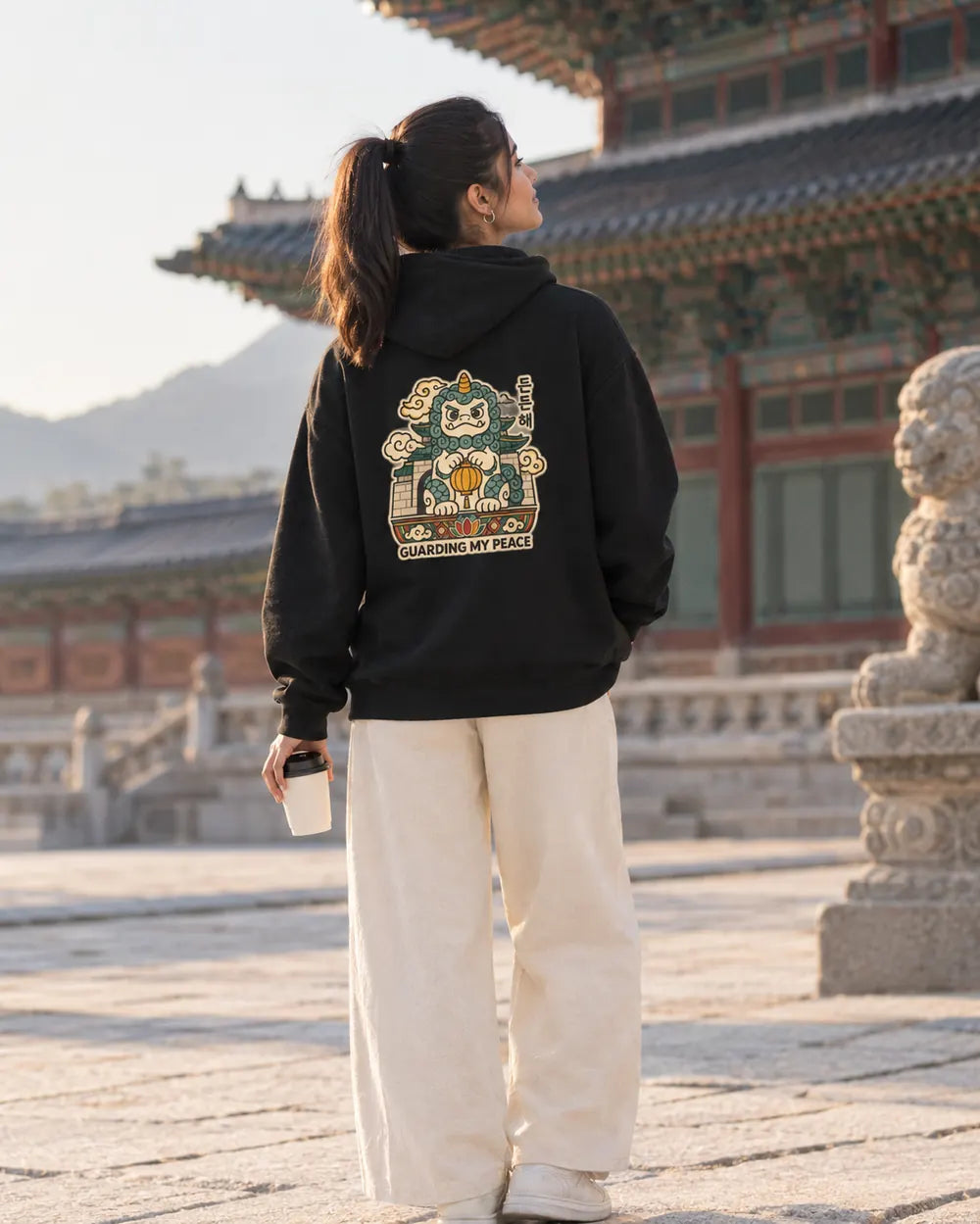 A woman stands with her back to the camera wearing a black hoodie with a large multicolor cartoon character graphic and the text 'GUARDING MY PEACE' on the back, paired with cream wide-leg pants, set against a traditional Korean palace courtyard with a stone lion statue.