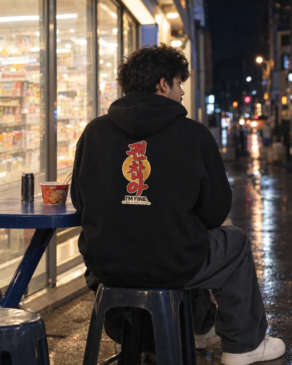 A person sitting on a stool at a blue table outside a convenience store at night, wearing a black hoodie with a large red and yellow graphic on the back and gray jeans.