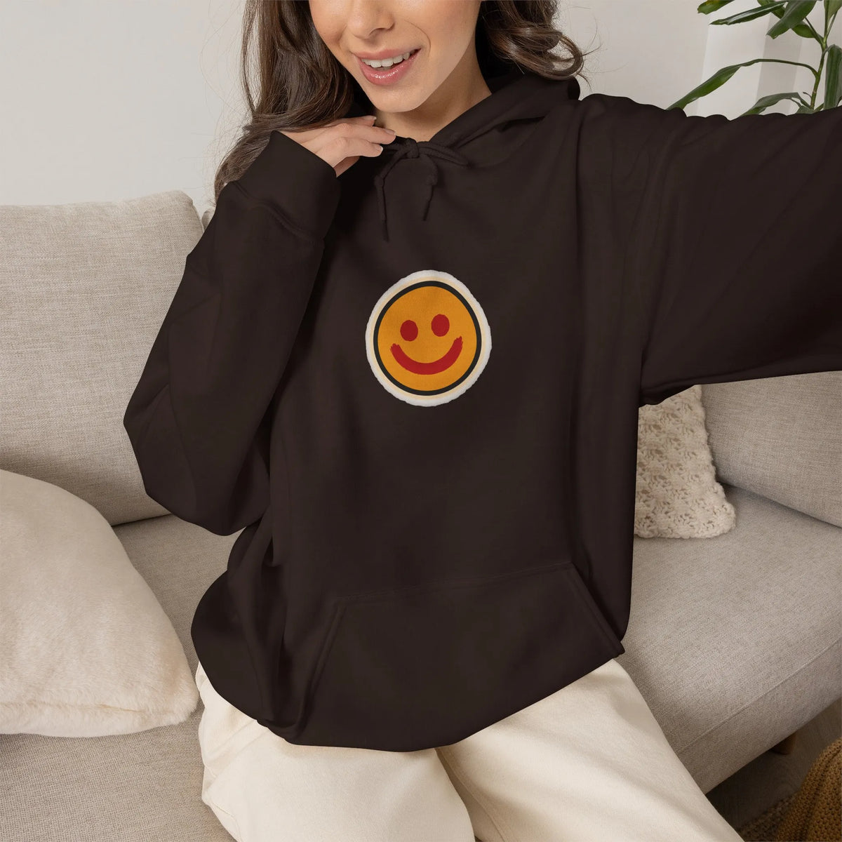 Woman on a sofa in a coffee brown hoodie with a small smiley face print on the chest