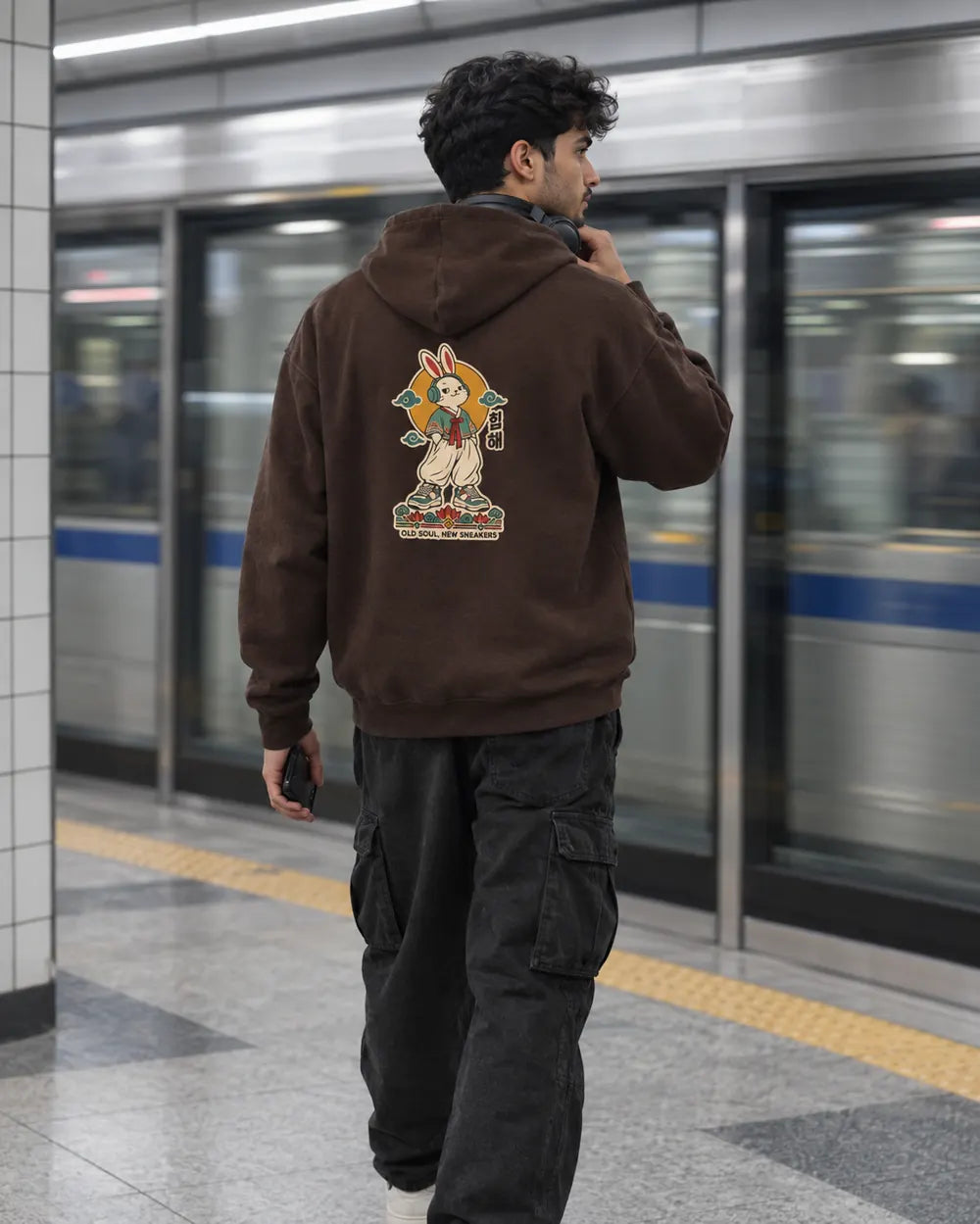 Man on a metro platform in a coffee brown Old Soul New Sneakers Korean hoodie and black cargo pants