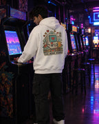 A man in a white hoodie with a retro computer graphic on the back stands in an arcade room, playing a game on a vintage arcade machine.