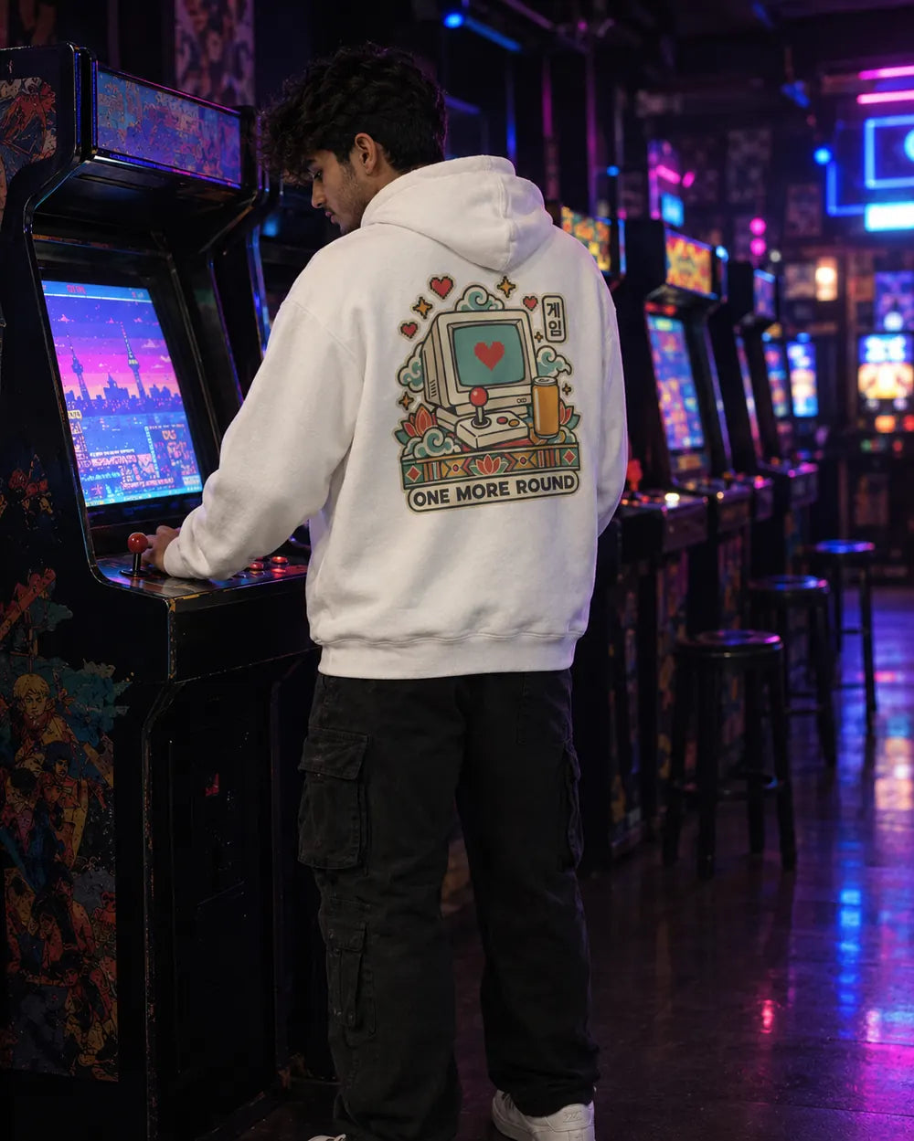 A man in a white hoodie with a retro computer graphic on the back stands in an arcade room, playing a game on a vintage arcade machine.