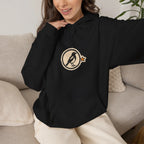 Woman relaxing on a sofa in a black hoodie with a small round athletic badge