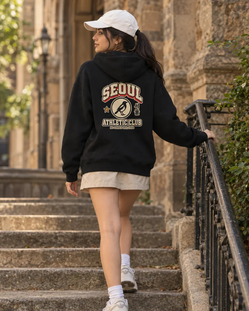 A woman wearing a black hoodie with a large 'SEOUL ATHLETIC CLUB' graphic on the back, a white cap, and a beige skirt walks down outdoor stone stairs next to a stone building.