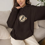 Woman on a sofa in a coffee brown hoodie with a small round athletic badge