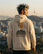 A man wearing a beige hoodie with a detailed graphic print on the back, standing outdoors with a blurred cityscape background at sunset.