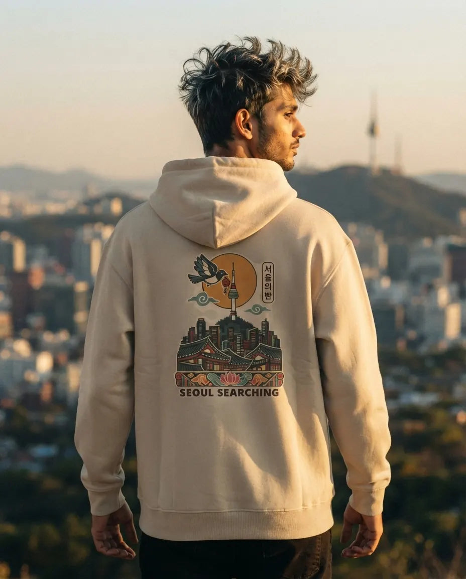 A man wearing a beige hoodie with a detailed graphic print on the back, standing outdoors with a blurred cityscape background at sunset.