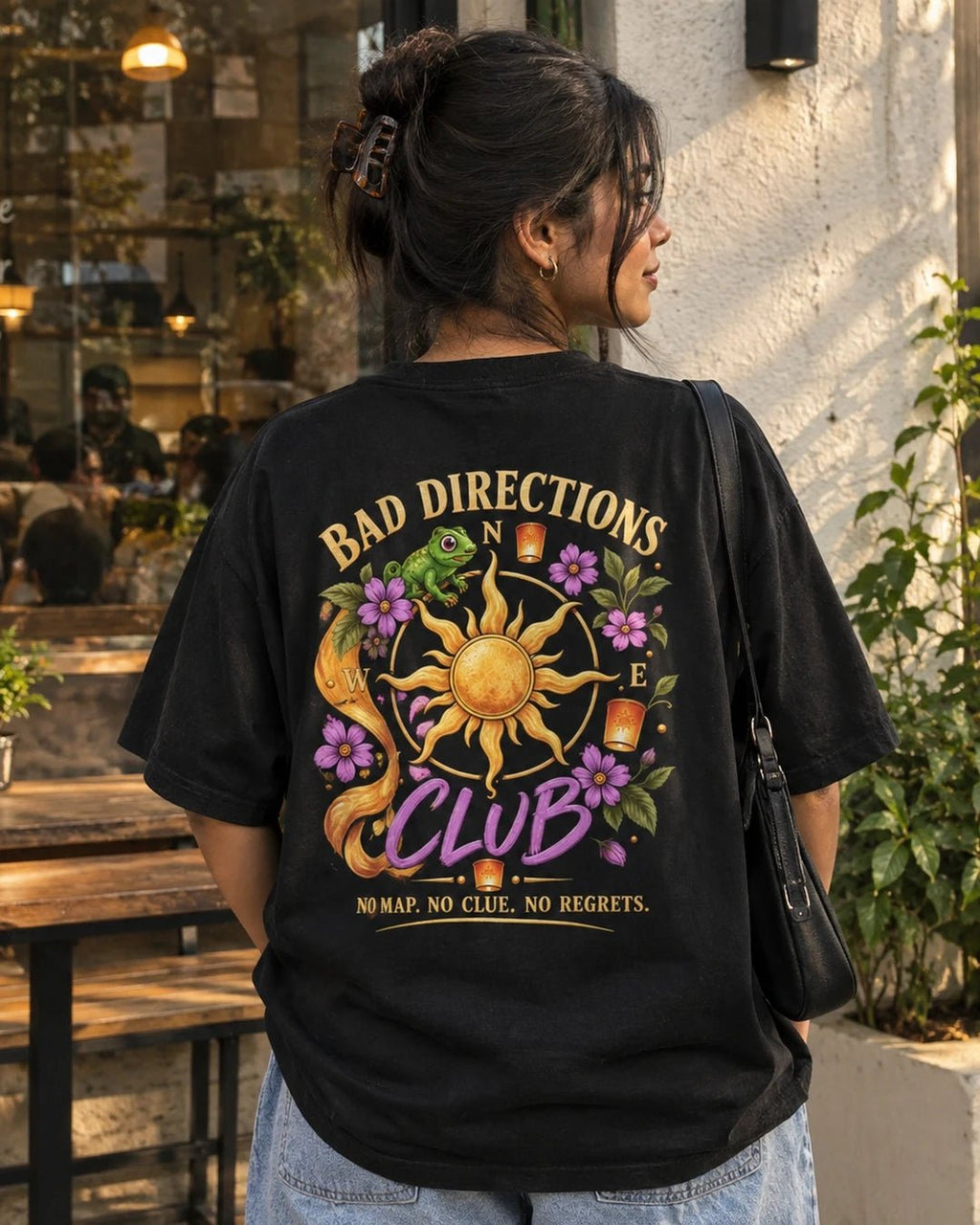 Woman wearing a black oversized Wear Pardesi t-shirt with a "Bad Directions Club" back print, a sun compass and purple flowers