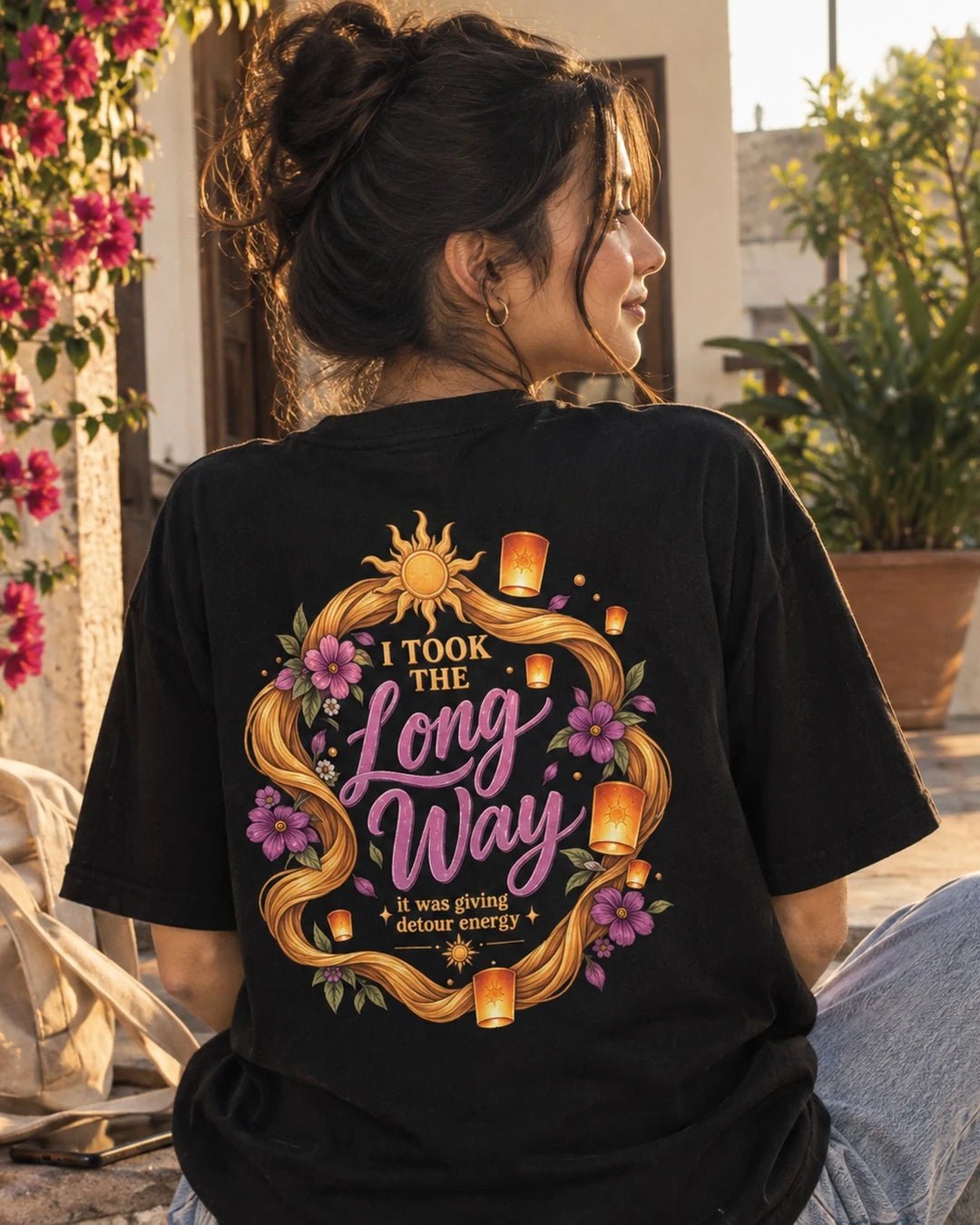 Woman wearing a black oversized Wear Pardesi t-shirt with an "I Took the Long Way" back print framed by golden braids and flowers